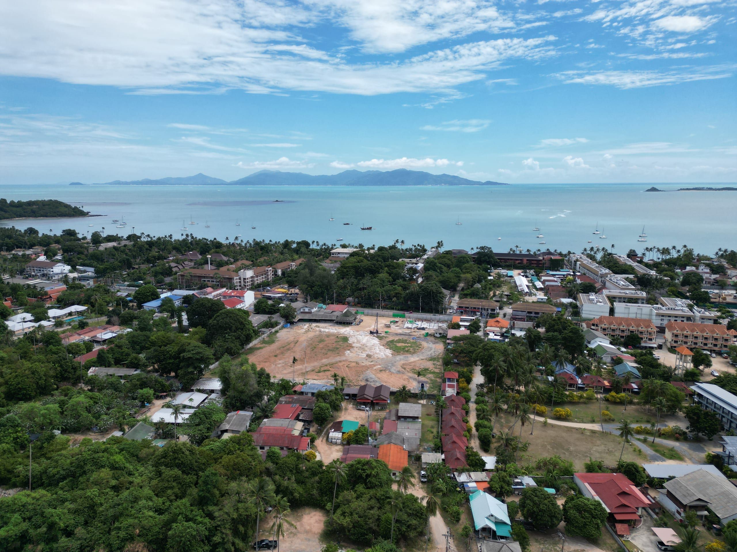Aerial view over a cleared plot among low-rise buildings and palms, with a bay, sailing boats and islands beyond