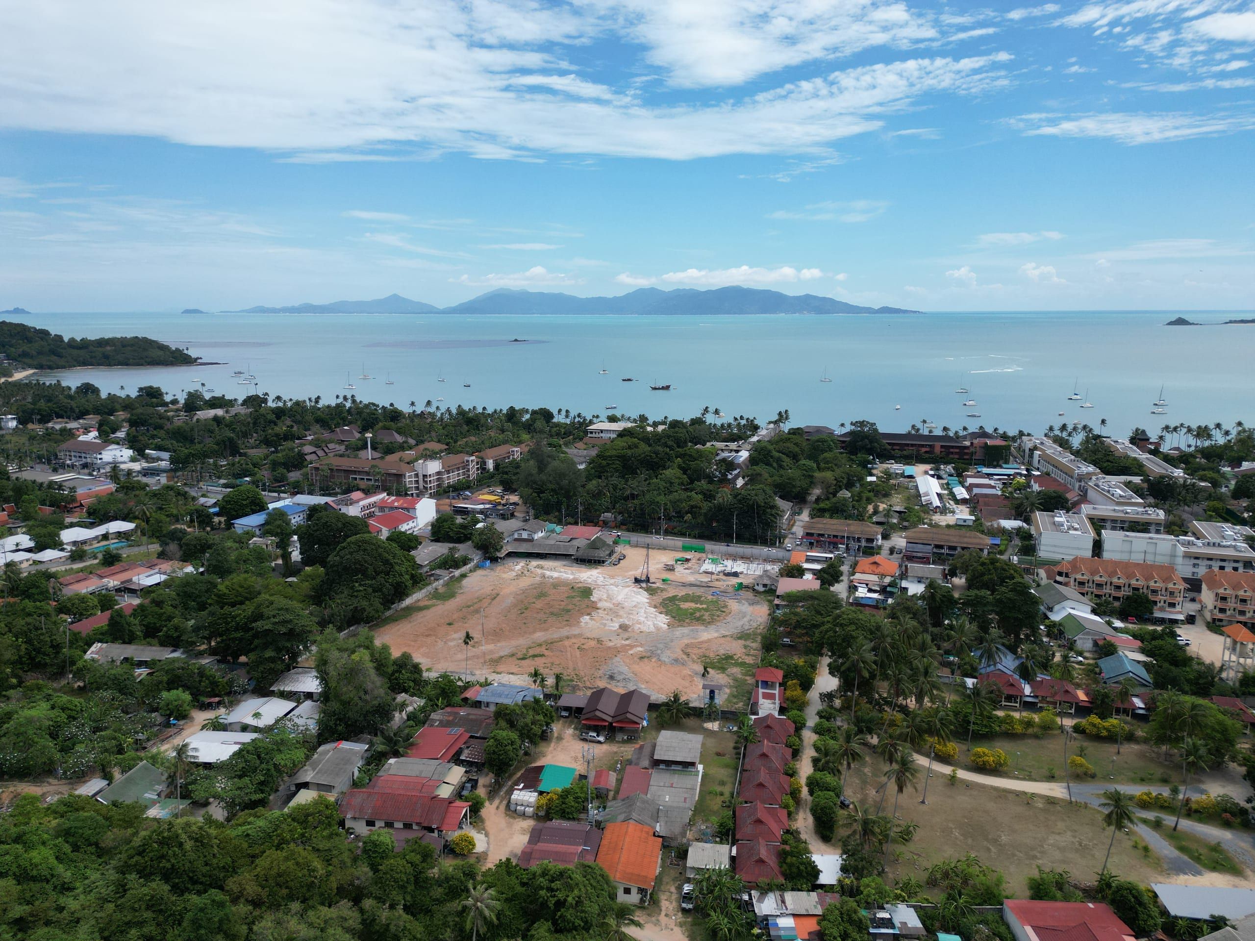 Aerial view of a cleared plot with a piling rig, ringed by low-rise buildings and trees, with the bay behind