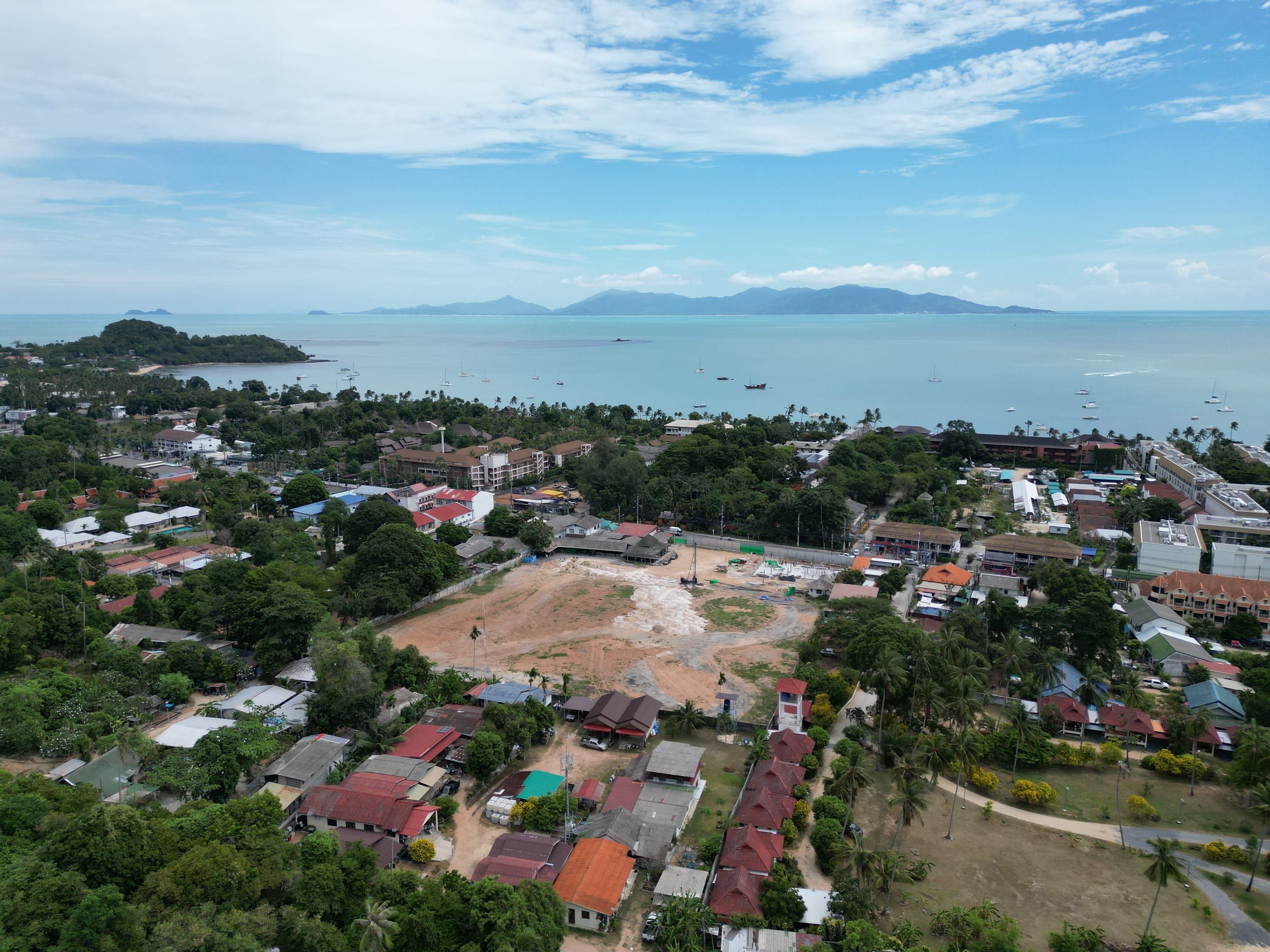 Aerial view of a cleared plot beside a green park-like area, with the bay, boats and a wooded headland behind