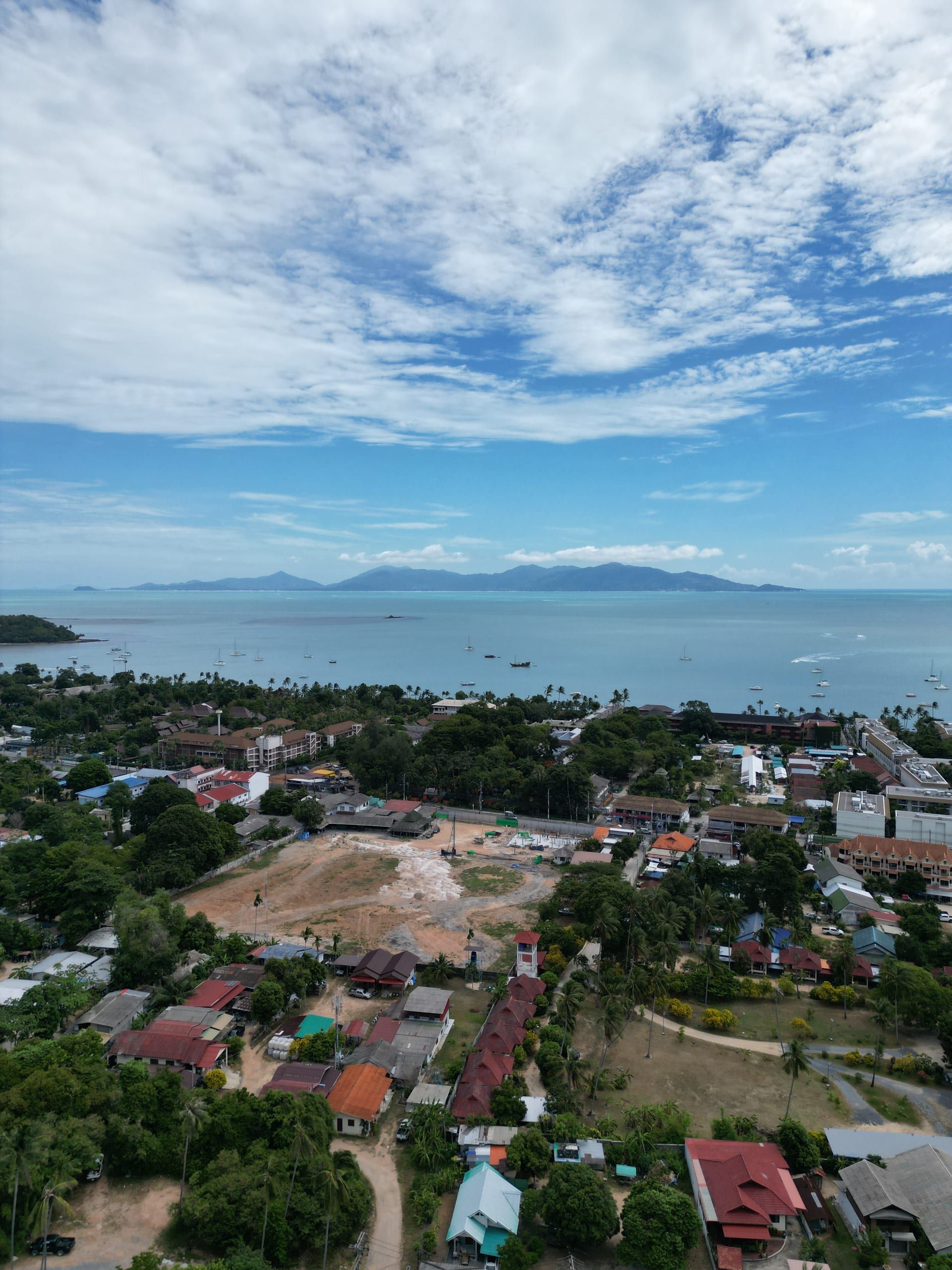 Aerial view over a cleared plot and low-rise rooftops towards a bay with moored yachts and distant hills