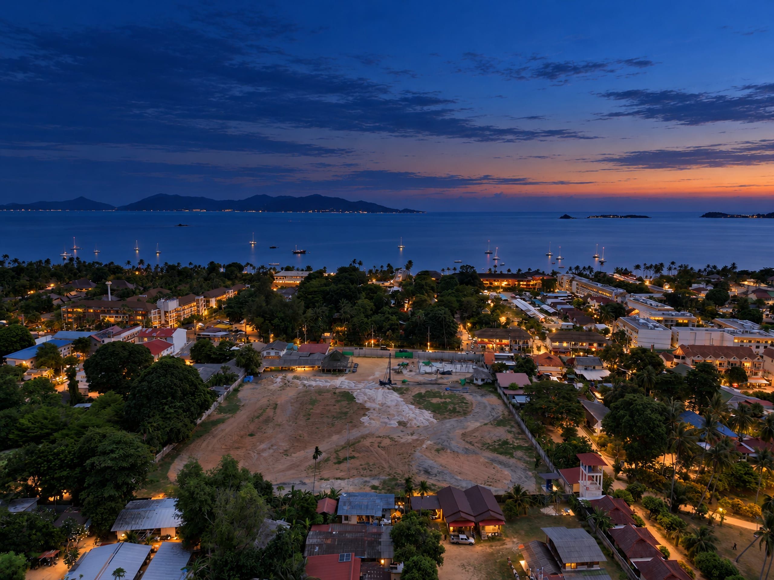 Wide aerial view of a cleared plot with a piling rig, low-rise streets around it and a calm bay with sailing boats
