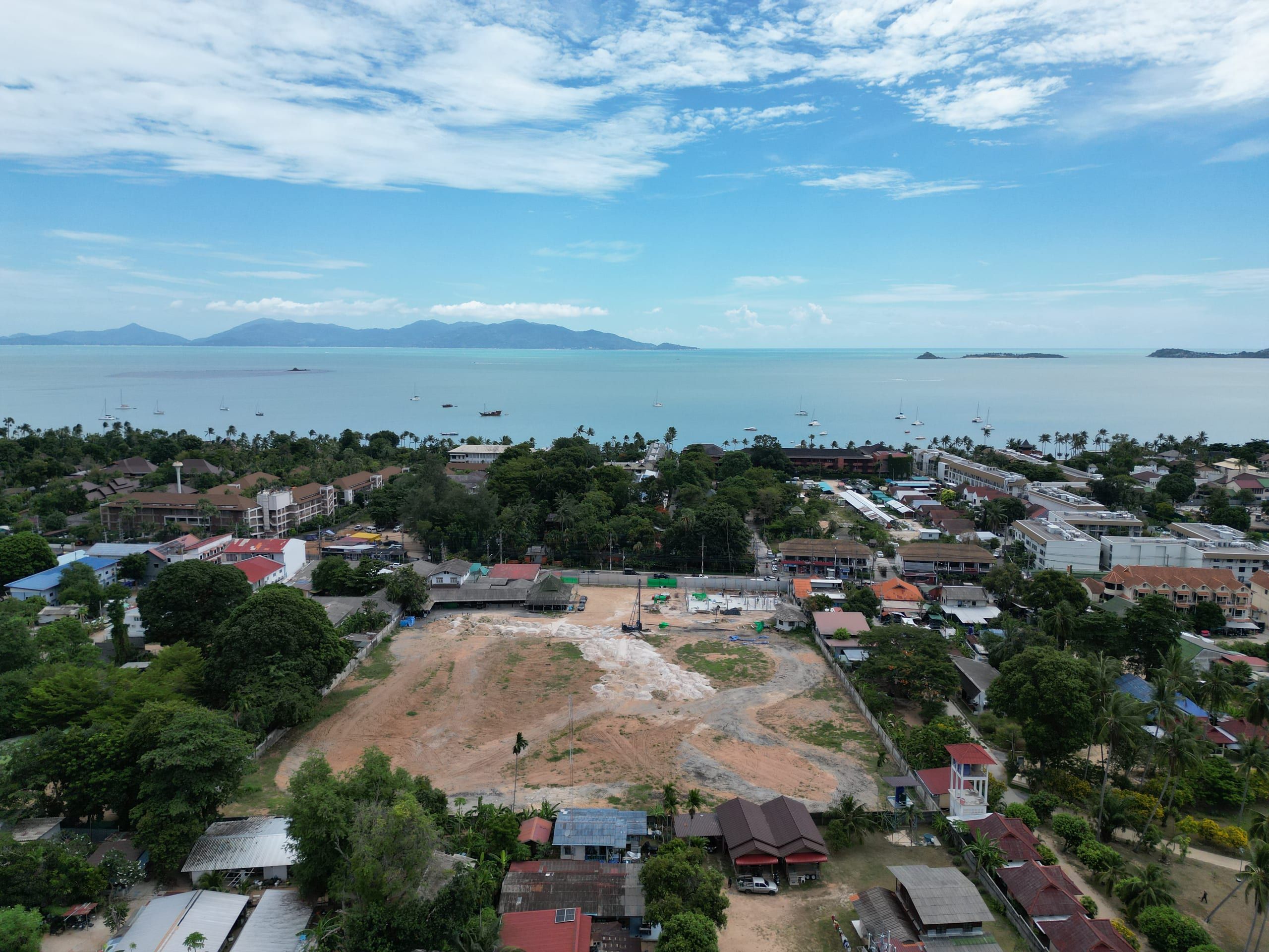 Wide aerial view of a cleared plot with a piling rig, low-rise streets around it and a calm bay with sailing boats