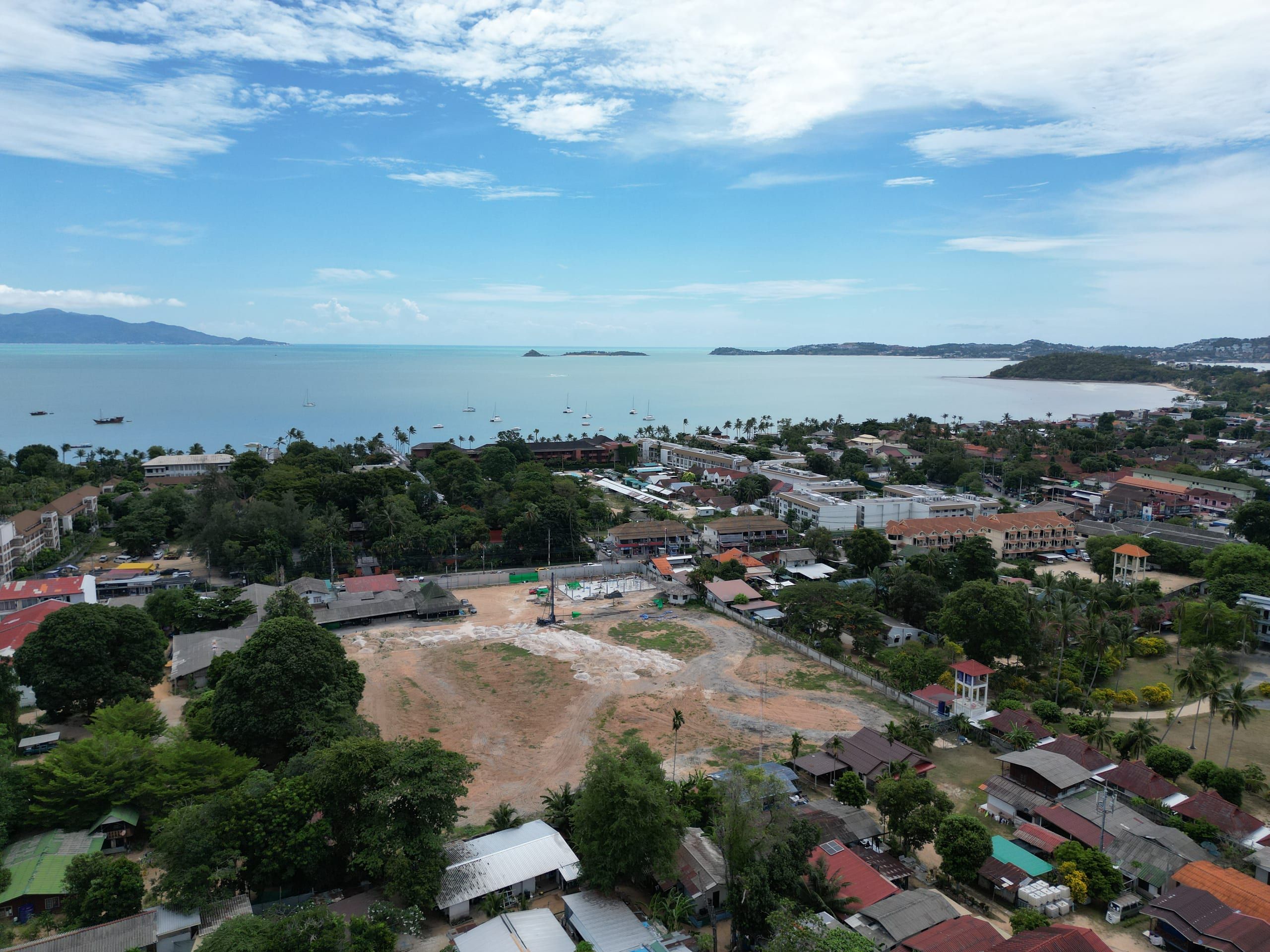 Aerial view of a cleared plot with a piling rig and early foundations, with houses, trees and a bay beyond