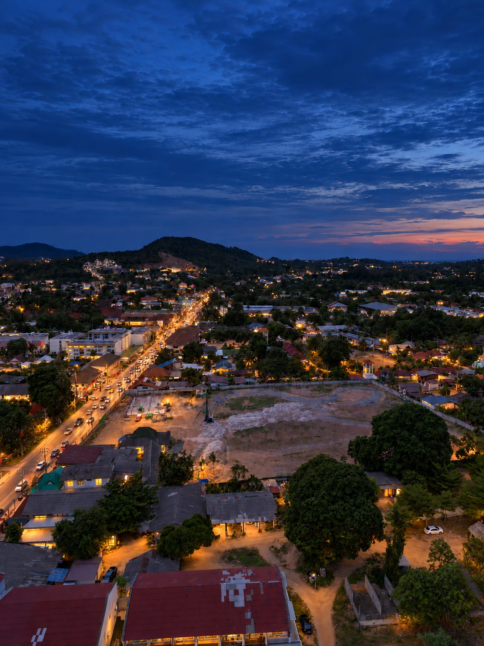 Aerial view of a cleared plot with a piling rig and first foundations beside a busy road, hills behind under cloud