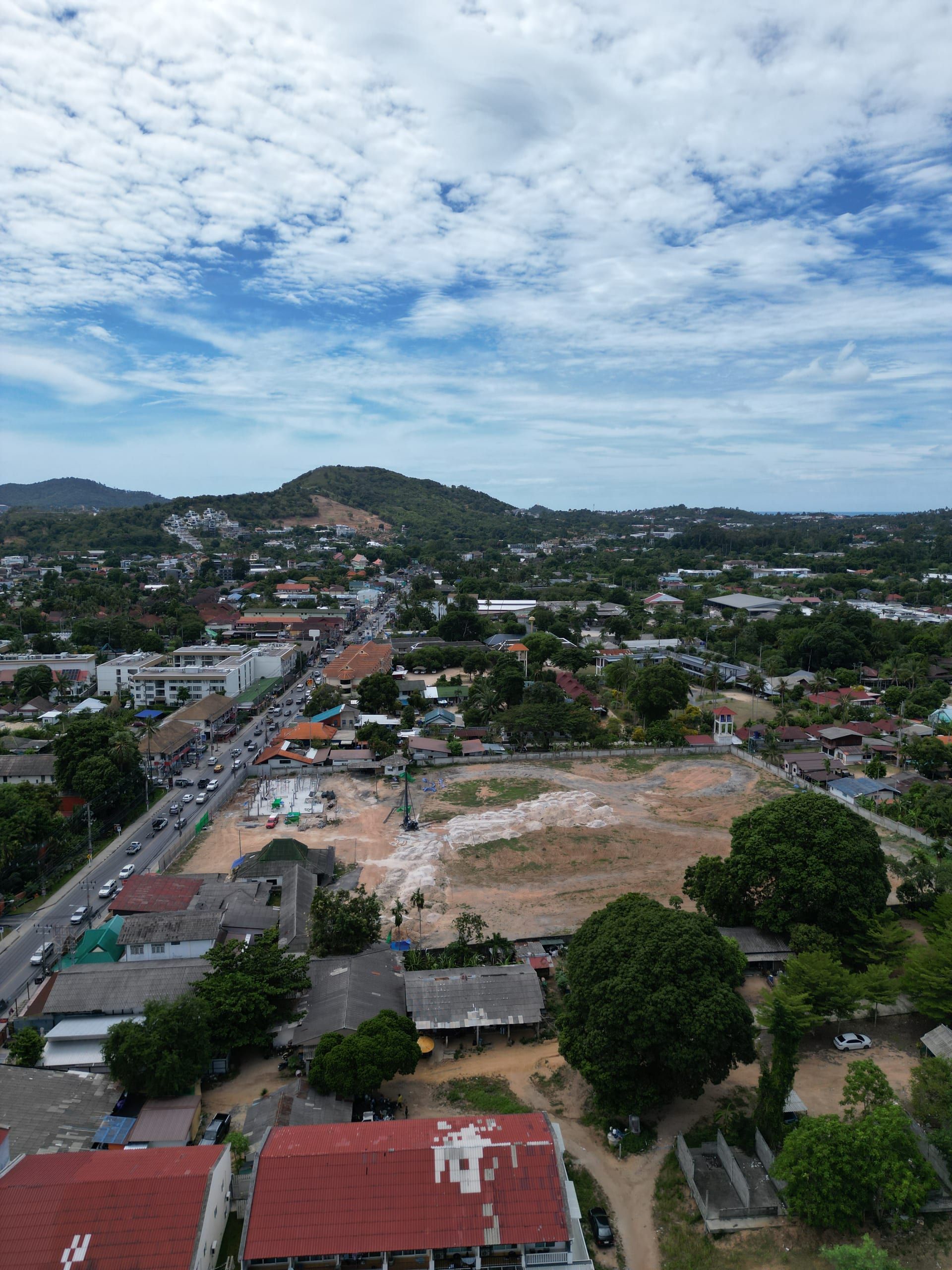 Aerial view of a cleared plot with a piling rig and first foundations beside a busy road, hills behind under cloud