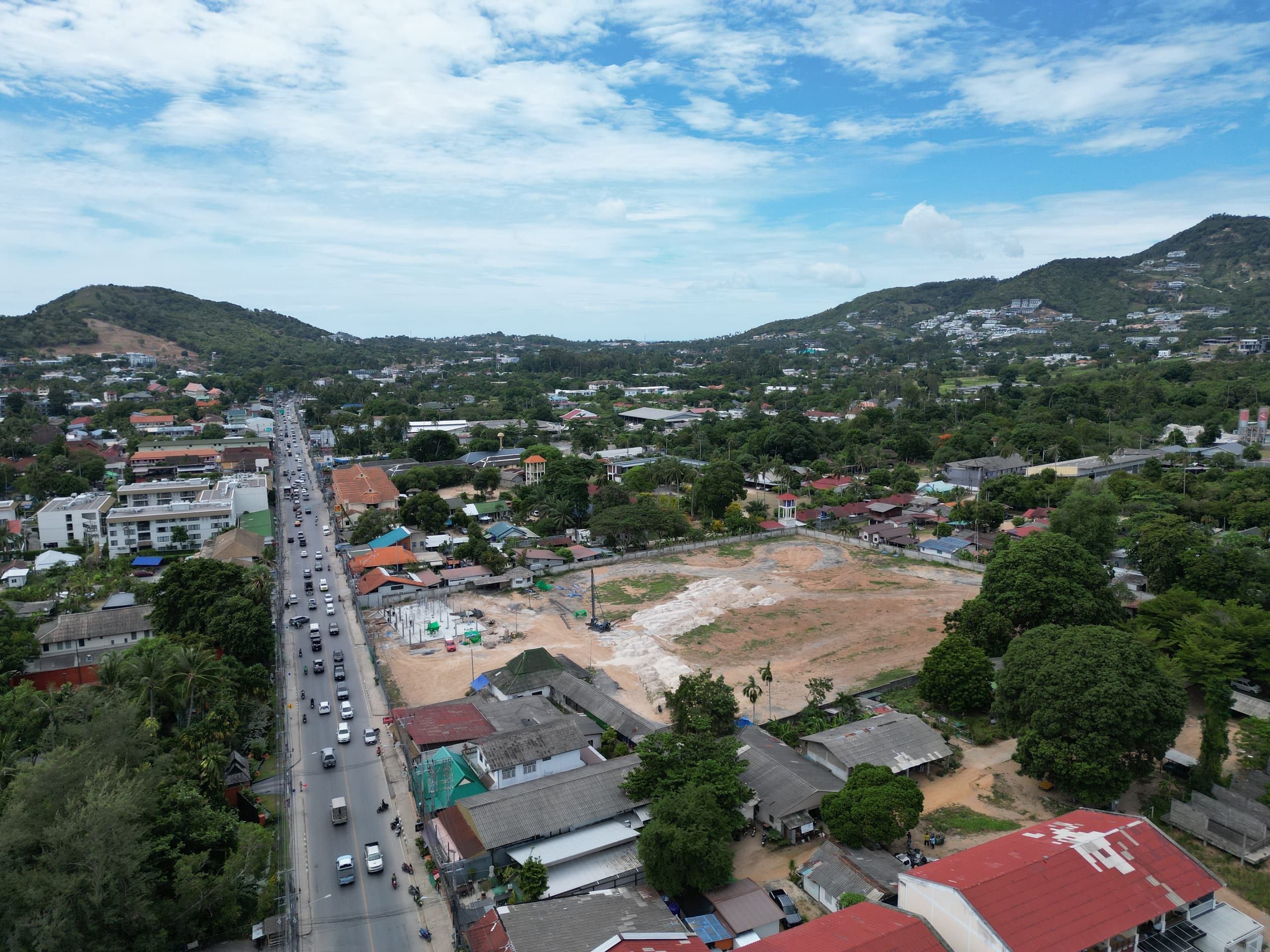 Aerial view along a busy road towards a cleared plot with a piling rig, with houses, trees and green hills behind