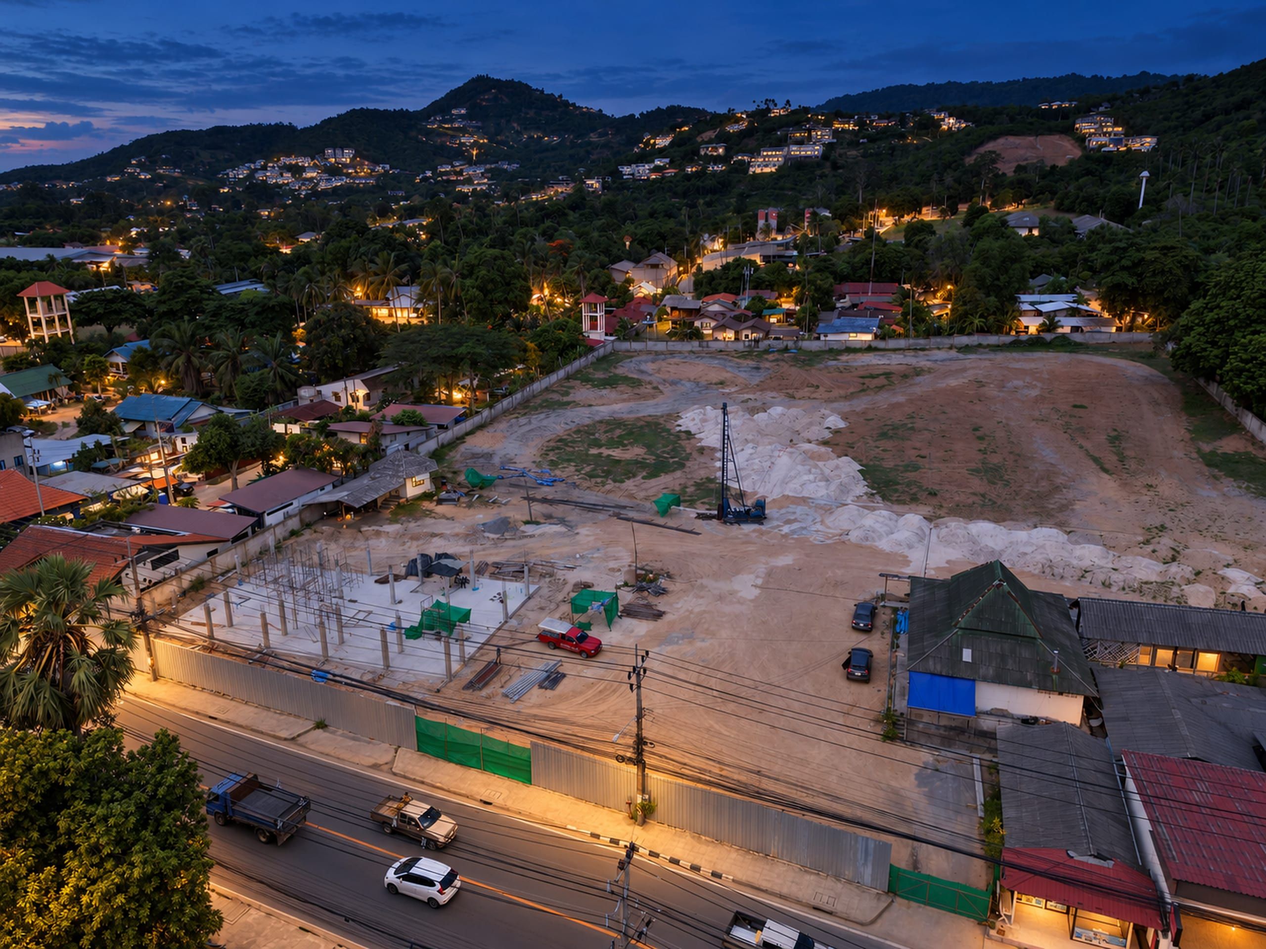 Close aerial view of the construction site: concrete slab with columns, piling rig, hoarding along the road and wooded hills