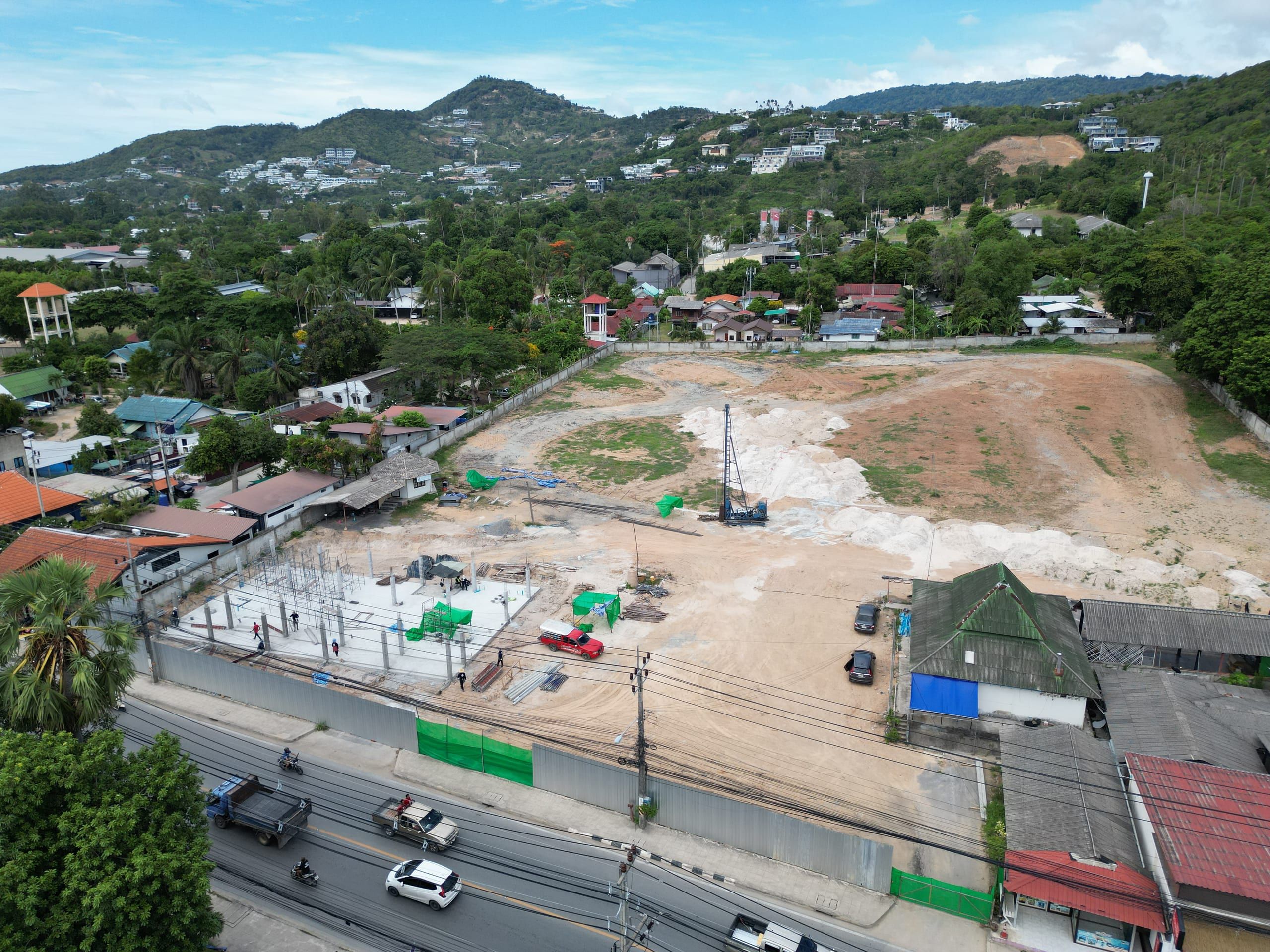 Close aerial view of the construction site: concrete slab with columns, piling rig, hoarding along the road and wooded hills