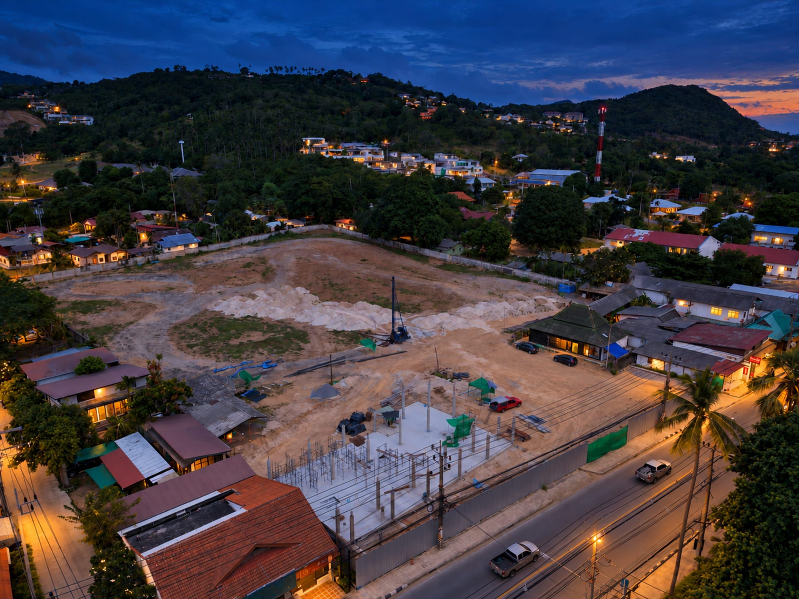 Aerial view of a walled plot with a piling rig and a concrete slab with columns, beside a road and with hills in the background