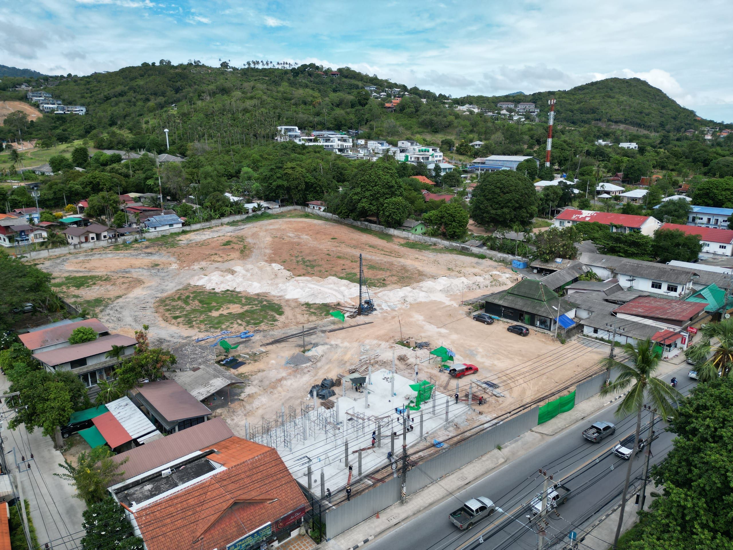 Aerial view of a walled plot with a piling rig and a concrete slab with columns, beside a road and with hills in the background