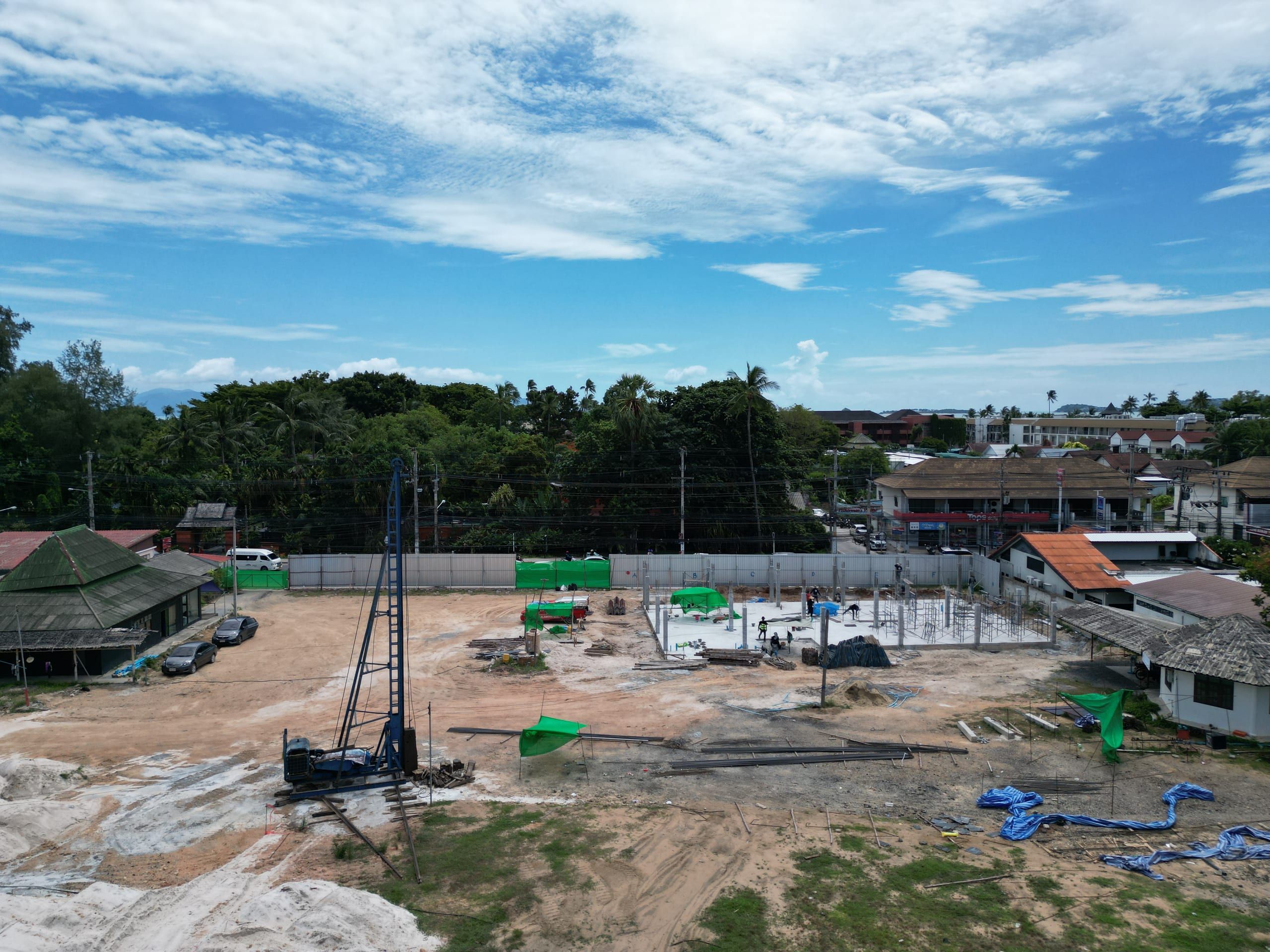 Aerial view of a building site with a piling rig, a concrete slab with columns, workers and neighbouring shops and trees
